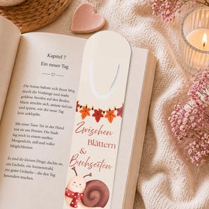 Puede incluir: Un marcapáginas color crema con temática otoñal. Presenta una guirnalda de hojas de otoño y un caracol de acuarela con una bufanda. El marcapáginas tiene el texto "Zwischen Blättern & Buchseiten".
