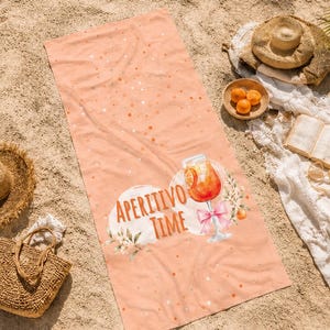 Könnte beinhalten: Ein pfirsichfarbenes Strandtuch mit der Aufschrift "APERITIVO TIME" und einer Aquarellillustration eines Cocktails. Die Szene umfasst einen Strohhut, eine Korbtasche, eine Schale mit Orangen und ein Buch, alles auf einem Sandstrand.