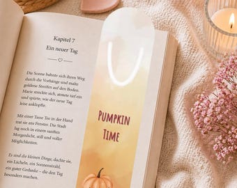 May include: A bookmark with the text "PUMPKIN TIME" and illustrations of pumpkins in shades of orange, white, and pink. The bookmark is placed in an open book with German text. The scene includes a candle, flowers, and a heart-shaped object.