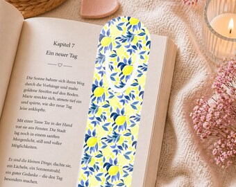 May include: A patterned bookmark with a lemon and leaf design in yellow and blue, placed in a book with German text. The image includes a lit candle, pink flowers, and a heart-shaped object. The bookmark is a reading accessory.