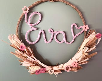 May include: A pink yarn wreath with the name "Eva" spelled out in pink yarn. The wreath is decorated with pink and white wooden flowers and natural straw.