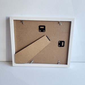 May include: White picture frame with a cardboard backing and metal hanging hardware. The frame is on a white surface.