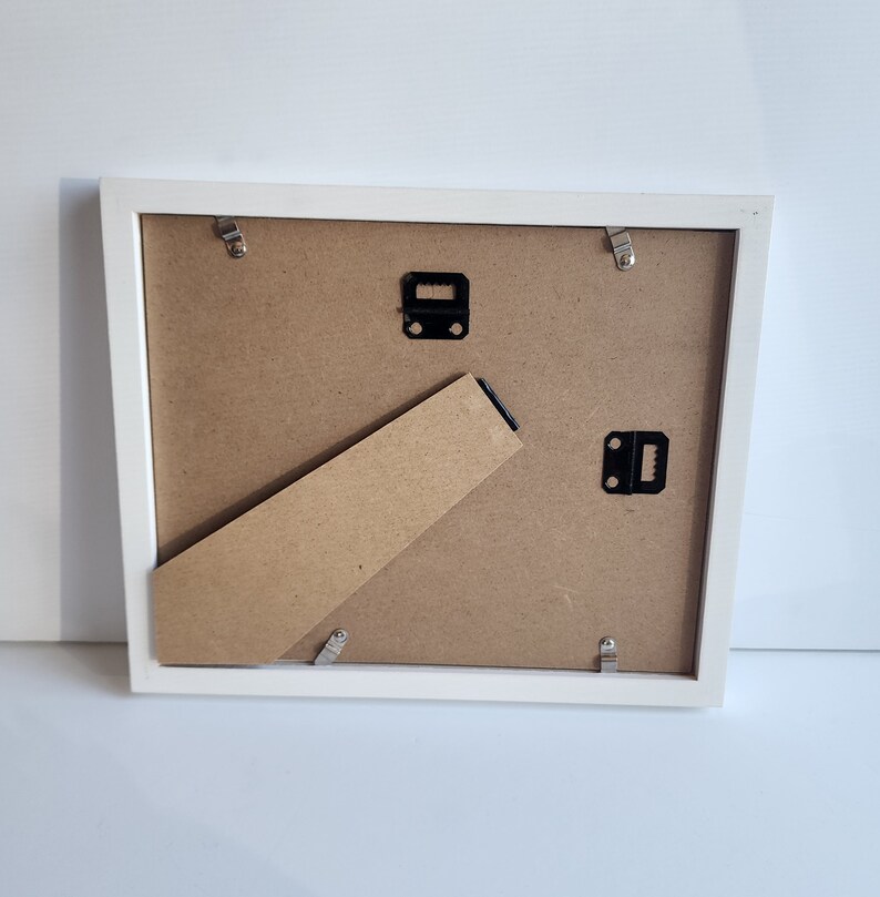 May include: White picture frame with a cardboard backing and metal hanging hardware. The frame is on a white surface.