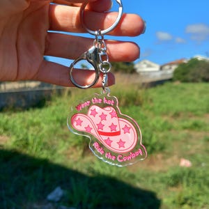 Könnte beinhalten: Transparenter Acryl-Schlüsselanhänger mit einem rosa Cowboyhut-Design und dem Text "Wear the hat, Ride the Cowboy" in Pink. Der Schlüsselanhänger hat einen silbernen Schlüsselring und Verschluss. Der Hintergrund ist ein blauer Himmel und grünes Gras.