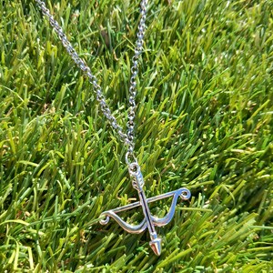 May include: Silver necklace with a bow and arrow pendant. The pendant is a detailed miniature of a bow and arrow. The necklace chain is a delicate link style. The jewelry is set against a backdrop of green grass.
