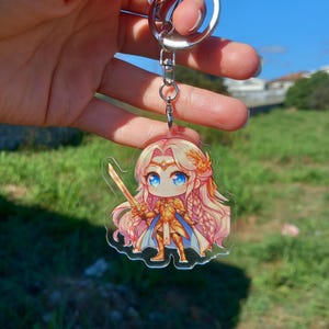May include: A clear acrylic keychain with a cartoon illustration of a character with long pink hair, blue eyes, and a golden sword. The character is wearing golden armour and a blue cape. The keychain has a silver metal ring.