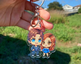 May include: A clear acrylic keyring featuring a cartoon couple. The male character is in a blue suit, and the female character is in a blue dress. The keyring has a silver ring and clasp. The couple has large eyes and detailed clothing.
