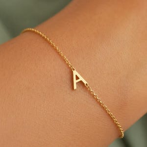 May include: A delicate gold-toned chain bracelet featuring a single capital letter A charm centered on the chain. The bracelet is constructed with fine circular links and a polished finish, creating a simple and refined piece of jewelry.