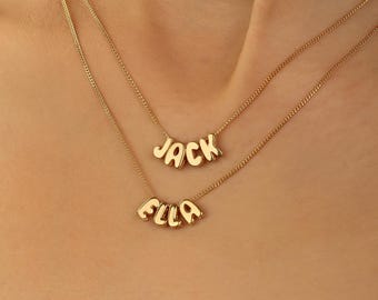 May include: Two gold-colored necklaces with delicate chains. The top necklace features the name "JACK" in bubble letters, and the bottom necklace displays the name "ELLA". The necklaces are worn around the neck.