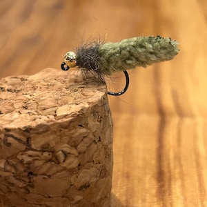 May include: A close-up of a handmade fly fishing lure. The lure features a gold bead head, a black hook, and a body made of olive green and brown materials. The lure is resting on a cork surface.