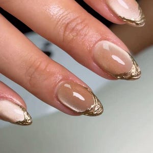 May include: Oval-shaped fingernails featuring a glossy, semi-sheer nude base coat with a subtle shimmer. The tips of the nails are adorned with a textured, metallic gold polish applied in a decorative, uneven line that follows the nail edge.