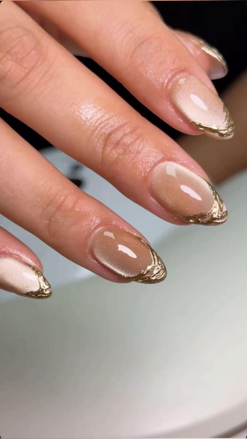 May include: Oval-shaped fingernails featuring a glossy, semi-sheer nude base coat with a subtle shimmer. The tips of the nails are adorned with a textured, metallic gold polish applied in a decorative, uneven line that follows the nail edge.