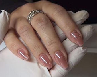May include: A hand with long, oval-shaped nails painted in a sheer, mauve color. The nails have a glossy finish. A silver ring with multiple rows of small, clear stones adorns the ring finger. The hand is wearing a white glove.
