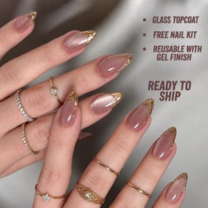 May include: Press-on nails with a mauve cat-eye effect and gold chrome tips. Text reads: GLASS TOPCOAT, FREE NAIL KIT, REUSABLE WITH GEL FINISH, READY TO SHIP. The nails are almond-shaped and worn with various gold rings featuring small clear stones.