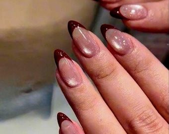 May include: Close-up of a hand with long, almond-shaped nails. The nails feature a classic French manicure design with a deep burgundy tip and a shimmering, light pink base. The nails are well-manicured and polished.