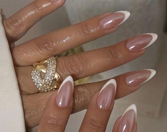 May include: Almond-shaped nails featuring a classic French manicure with a glossy nude base and crisp white tips. A large, gold-toned statement ring with a textured surface and embedded clear crystals is worn on the ring finger of the hand.