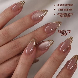 May include: Almond-shaped press-on nails in a soft nude pink with a shimmering cat-eye effect and gold foil tips. Text reads: GLASS TOPCOAT, FREE NAIL KIT, REUSABLE WITH GEL FINISH, READY TO SHIP. These reusable nails feature a glossy gel finish.