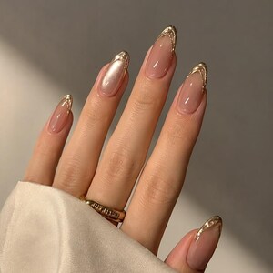 May include: Almond-shaped nails featuring a sheer nude base color with metallic gold tips. The nails have a glossy finish and a smooth, rounded edge. A gold band ring with a textured surface is worn on the ring finger, complementing the gold nail art.