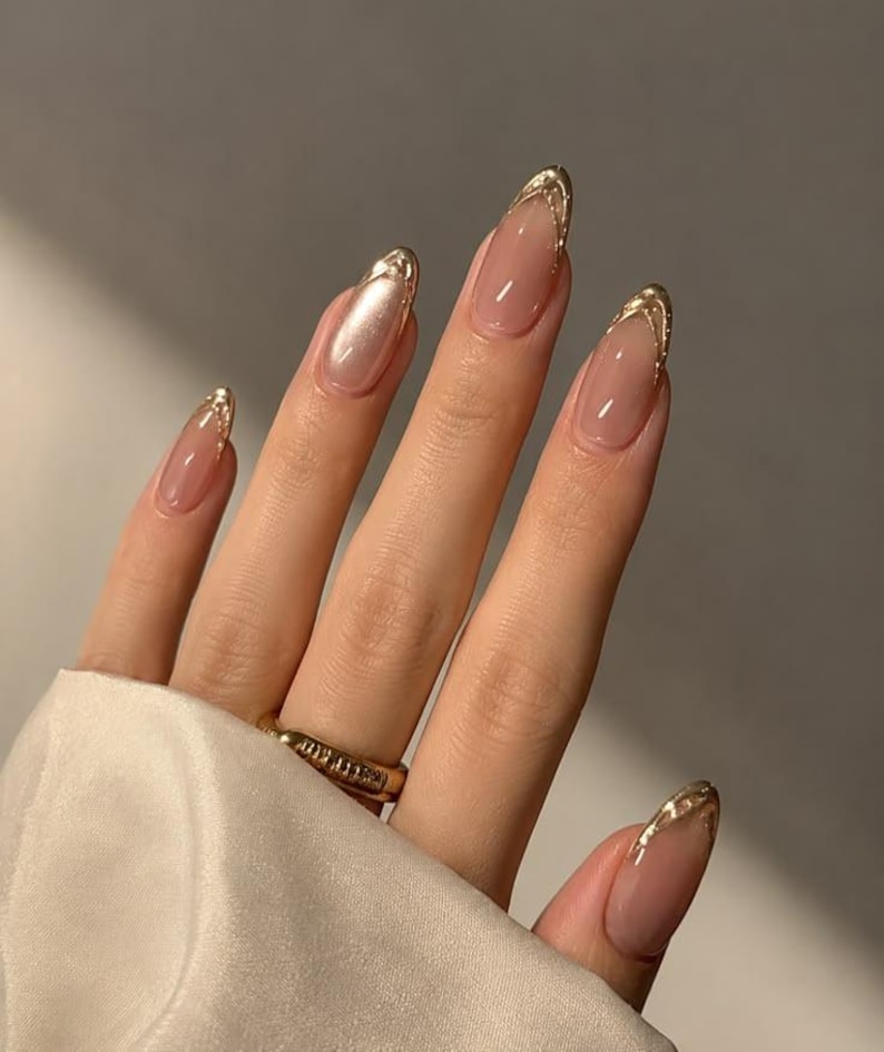 May include: Almond-shaped nails featuring a sheer nude base color with metallic gold tips. The nails have a glossy finish and a smooth, rounded edge. A gold band ring with a textured surface is worn on the ring finger, complementing the gold nail art.