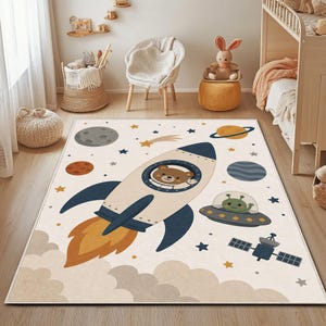 May include: A rectangular area rug featuring a space-themed design on a cream background. The print includes a rocket ship piloted by a brown bear, a green alien in a flying saucer, a satellite, planets, stars, and clouds in shades of blue and orange.