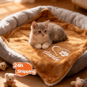Può includere: Un gattino grigio e marrone riposa su un letto per animali domestici marrone e morbido con una coperta soffice. La coperta è ricamata con un cuore, un'impronta di zampa e il nome "Annie". Il letto ha un bordo grigio e bianco.