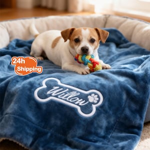 May include: A small dog with brown and white fur lies on a blue blanket, holding a colourful rope toy. The blanket has a bone-shaped design with the name "Willow" embroidered in white. The dog is in a dog bed.
