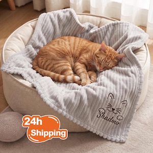 Peut inclure: Un panier pour animal de compagnie rond et crème contenant une couverture en polaire côtelée gris clair. Un chat tigré orange est recroquevillé et endormi sur la couverture. Elle présente un motif brodé de visage de chat et le nom Shadow.