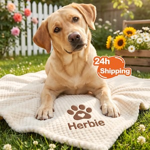 Puede incluir: Una manta texturizada de color crema con una huella de pata marrón bordada, un pequeño corazón en el centro y el nombre Herbie escrito debajo. Un perro Labrador retriever amarillo está descansando sobre la manta al aire libre.
