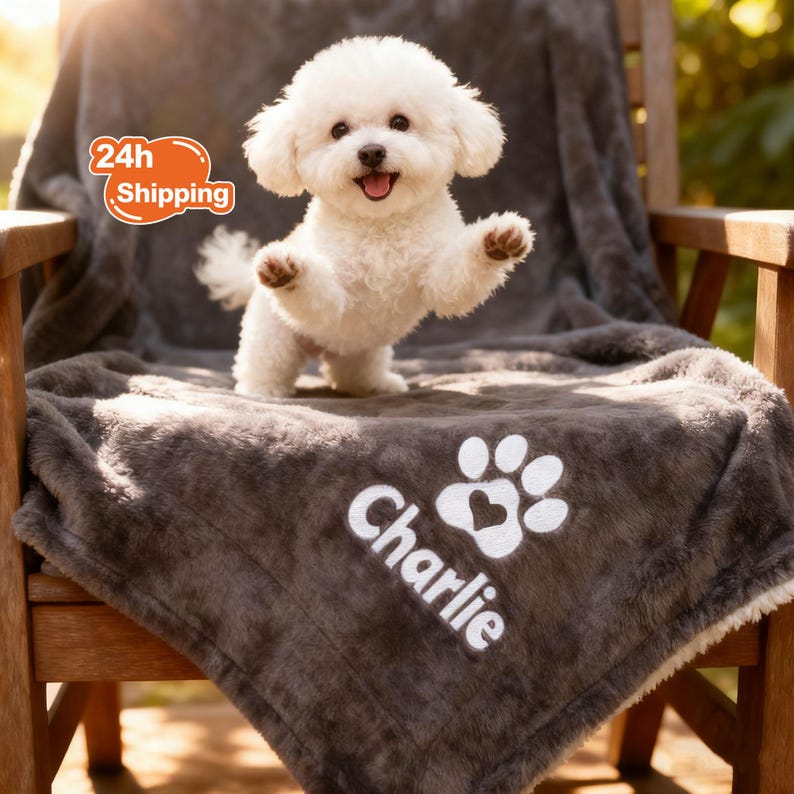 May include: A fluffy white Bichon Frise dog stands on a gray blanket with the name "Charlie" and a paw print design. The blanket is draped over a wooden chair. The dog is smiling with its paws raised.