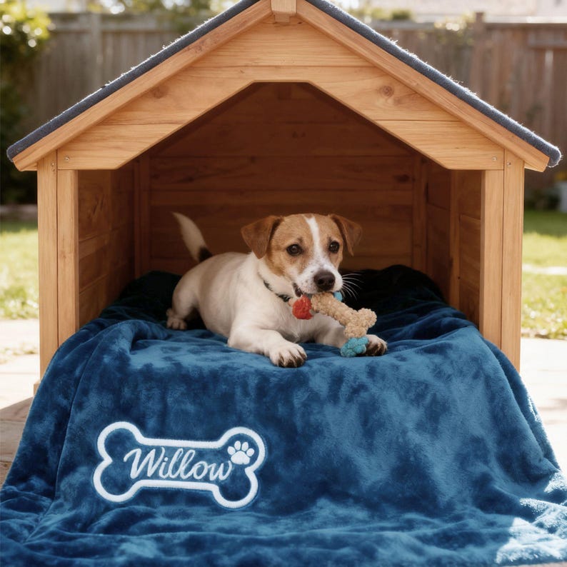 May include: A small dog rests inside a wooden dog house, lying on a blue blanket. The blanket has the name "Willow" embroidered on it in white. The dog is holding a bone-shaped toy.