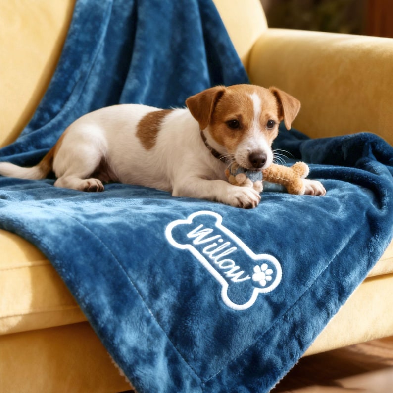May include: A Jack Russell Terrier puppy lies on a blue blanket embroidered with the name "Willow" in white. The puppy is white with brown markings and holds a small toy. The blanket is on a yellow armchair.