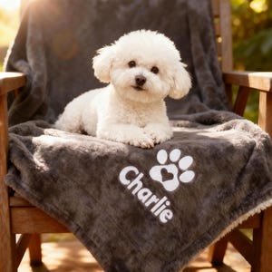May include: A fluffy white Bichon Frise dog rests on a gray, plush blanket draped over a wooden chair. The blanket is personalized with the name "Charlie" and a paw print design in white.