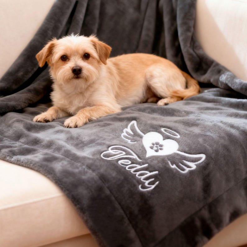 May include: A small, tan dog rests on a gray blanket. The blanket has a white embroidered design of a heart with wings, a paw print, and the name "Teddy". The blanket is draped over a light-colored surface.
