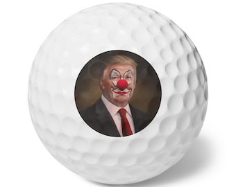 May include: A white golf ball with a round sticker featuring a portrait of a man with a red clown nose. The man is wearing a dark suit and a red tie.