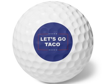 May include: A white golf ball with a dimpled surface. A blue circle with the text "LET'S GO TACO" in white letters is printed on the ball, framed by a red rectangle. The golf ball is designed for recreational use.