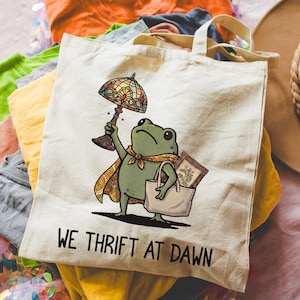 May include: A cream-colored canvas tote bag featuring a graphic illustration of a green frog wearing a patterned cape. The frog holds a stained-glass lamp and carries a smaller bag containing a framed picture. The text reads WE THRIFT AT DAWN.