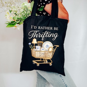 May include: Black tote bag with the text "I'd Rather Be Thrifting" in a script font. The bag features a gold shopping cart graphic filled with vintage items like a lamp, teacups, and plates. The bag is held by a person.
