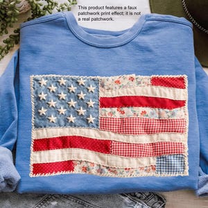 Könnte beinhalten: Ein hellblaues Sweatshirt mit Rundhalsausschnitt und einem zentralen rechteckigen Aufdruck einer amerikanischen Flagge mit Patchwork-Optik. Das Design umfasst verschiedene Muster wie rot-weiße Streifen, florale Drucke und weiße Sterne.