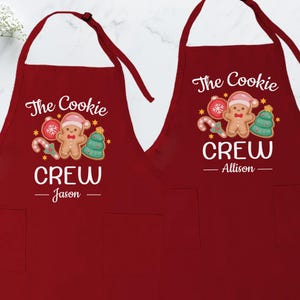 May include: Two red aprons with white text and gingerbread man designs. The text reads "The Cookie Crew" with names below. Each apron has a pocket and adjustable neck strap. Festive holiday kitchen apparel.