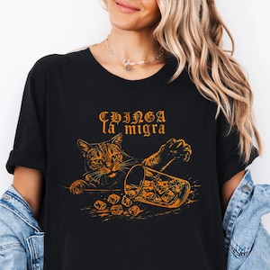 May include: Black t-shirt with an orange graphic of a cat reaching for a tipped-over glass of ice cubes. The text "CHINGA la migra" is above the cat. The shirt is a casual style.
