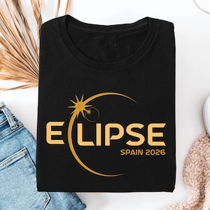 May include: A black t-shirt featuring a gold graphic of a solar eclipse. The design includes the text "ECLIPSE" with a stylized sun icon replacing the letter C, and the text "SPAIN 2026" printed in gold lettering below the main graphic.