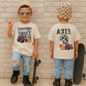 May include: Beige t-shirt with the text "Crushing Into Three" and a monster truck graphic. The back of the shirt has the name "Axel" and a number 3 with a monster truck graphic. The child is wearing blue jeans and a checkered hat.