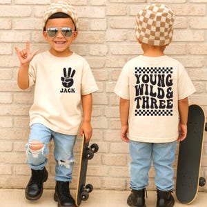 May include: A child wearing a beige t-shirt with a black hand symbol and the name "Jack". The child is also wearing light blue ripped jeans, black boots, and a checkered hat. The child is holding a skateboard.