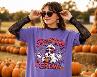 May include: A purple t-shirt featuring a graphic print of ghosts wearing sunglasses and a captain's hat, holding a tropical drink. The text reads SPOOOKY CRUISE CREW, accompanied by illustrations of a cruise ship, anchors, and pumpkins.