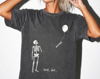 May include: A charcoal gray short-sleeve t-shirt featuring a white graphic of a skeleton looking up at a detached skeletal hand holding a white balloon. The text "Well, shit..." is printed in white below the skeleton on the front of the shirt.