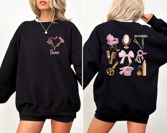 Op de afbeelding: Zwart sweatshirt met witte kraag met een roze hartontwerp met salon gereedschap en de naam "Jane". De achterkant van het sweatshirt toont verschillende illustraties gerelateerd aan salons, waaronder een stoel, spiegel en haarstyling tools.