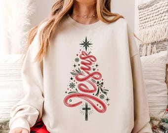 May include: A beige crewneck sweatshirt featuring a graphic design of a Christmas tree formed by the red cursive word Jesus. The tree is decorated with dark green pine branches, small snowflakes, stars, and a black cross at the base.