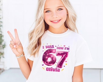 May include: A white t-shirt with the text "I WAS NOW I'M 67 Olivia" in pink and purple. The shirt is worn by a young girl with blonde hair styled in two buns, making a peace sign with her fingers.