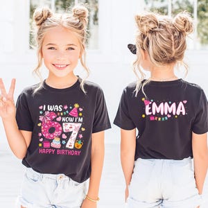 Pode incluir: T-shirt preto com o texto "I WAS 6 NOW I'M 7 HAPPY BIRTHDAY" e um gráfico colorido. A parte de trás da camisa tem o nome "EMMA" em rosa com um chapéu de festa e um design de banner. Um artigo de vestuário com tema de aniversário.