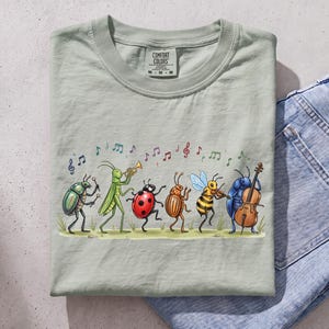 Peut inclure: Un t-shirt vert sauge clair orné d'un motif représentant six insectes jouant des instruments de musique, dont un scarabée avec des tambours, une sauterelle avec une trompette, une coccinelle, un doryphore, une abeille avec un violon et un scarabée bleu avec un violoncelle.